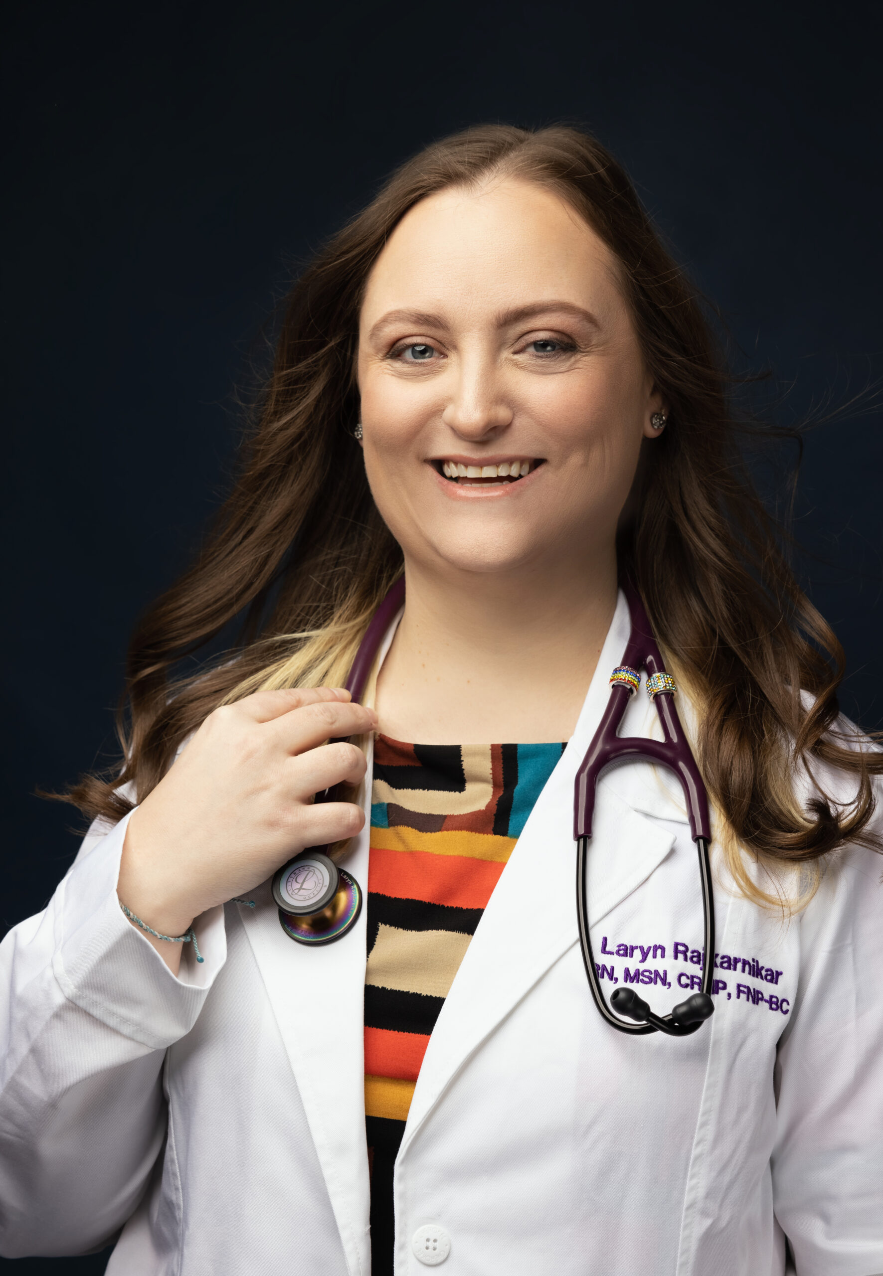 Laryn Rajnikarnikar is a smiling woman wearing a white lab coat over a striped shirt. She has dark hair and a stethoscope slung over her neck.