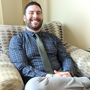 Stephan hoschar is a smiling man with short dark hair and a well trimmed beard. He is sitting in a chair wearing a blue plaid shirt, khaki pants and a green tie.