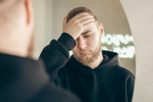 a man holds his hand to his forehead while standing in front of a mirror and wondering about treatment options for bipolar disorder