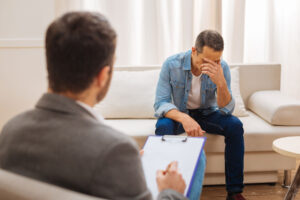 a man sits on a couch appearing to be struggling and holds his head down resting on his hand while his therapist sits across from him with notepad explaining options for anxiety treatment in meadowlands pennsylvania
