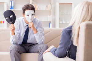 a man sits on a couch and holds up a white face mask to his face as he talks to his therapist about his narcissistic behaviors
