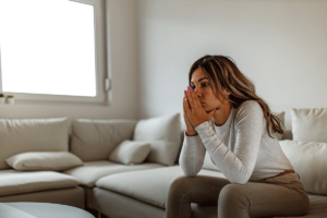 upset woman holds her hands to her face while sitting on a couch and thinks about self-harm prevention techniques