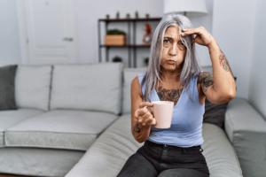 woman sits on a couch holding a cup of coffee in one hand and other hand to her head and wondering how does caffeine affect adhd