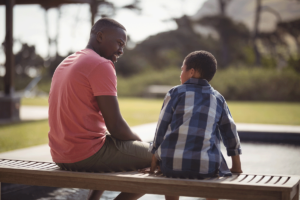 a father and young son sit on a bench talking as an example of why it's important to learn about talking about mental health with your kids