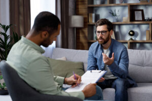 man with glasses sits on couch talking to a therapist while participating in therapy services at the greensburg office