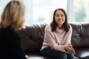 smiling woman sits on a couch and participating in psychiatric care with her psychiatrist sitting across from her